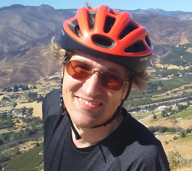Nathan on a mountain outside Wenatchee, WA wearing an orange mountain biking helmet, black shirt, and brown sunglasses.